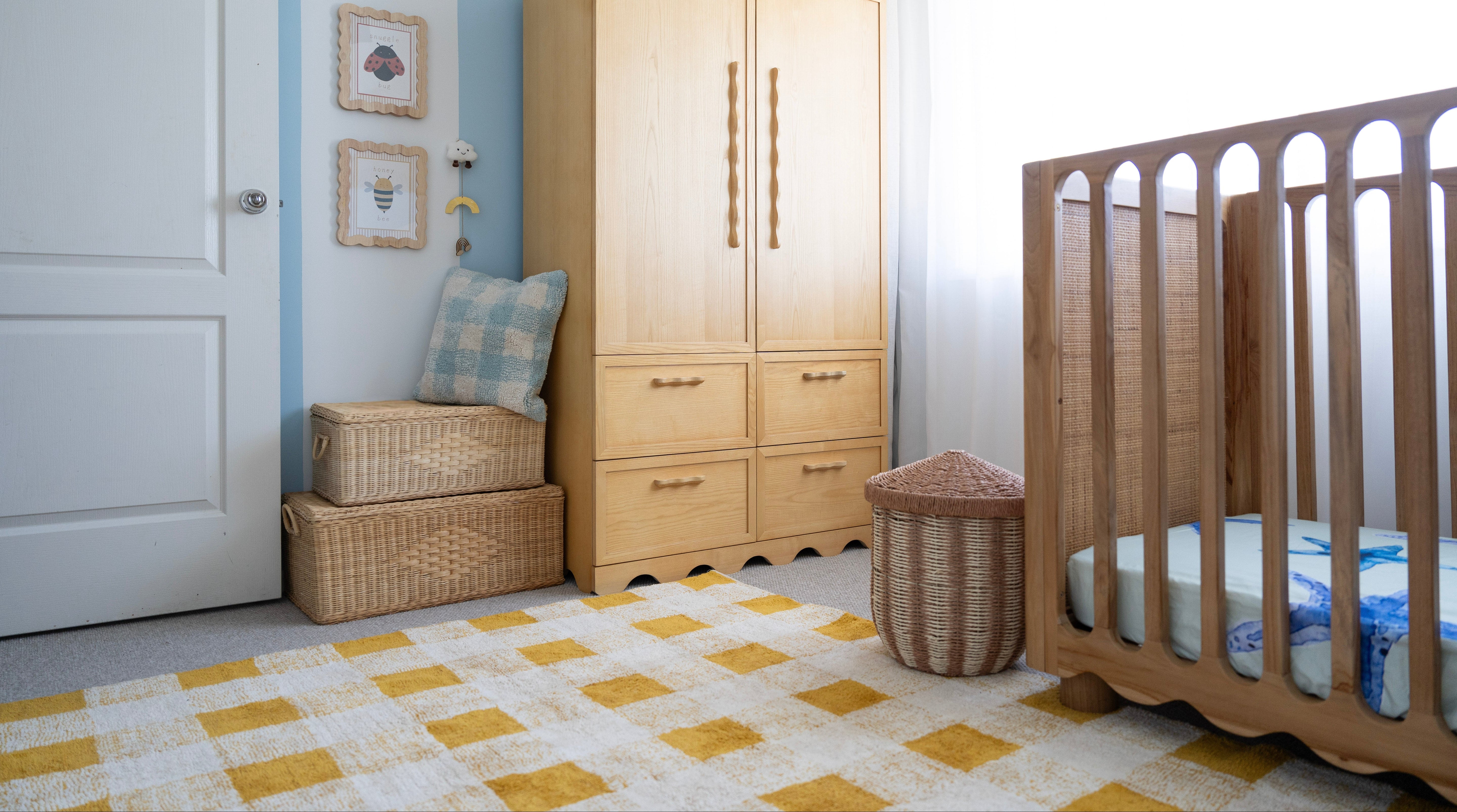 A styled nursery room featuring a light natural wood wardrobe, a wooden crib, a yellow and white checkered rug, and a blue and beige patterned pillow on a wicker bench.