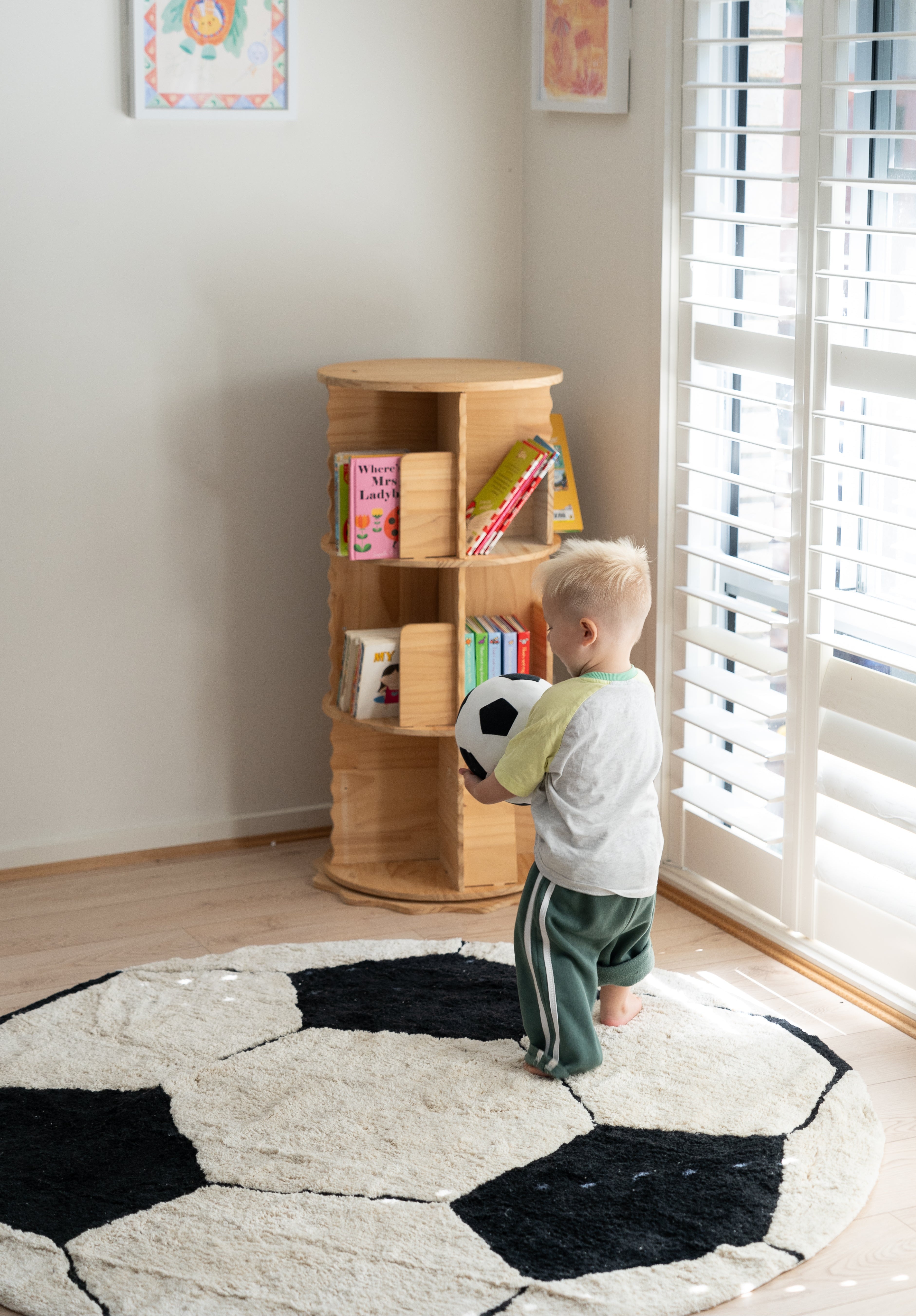 A toddler stands on a black-and-white soccer-ball patterned rug in a bright room, holding a soccer ball next to a small wooden bookshelf.