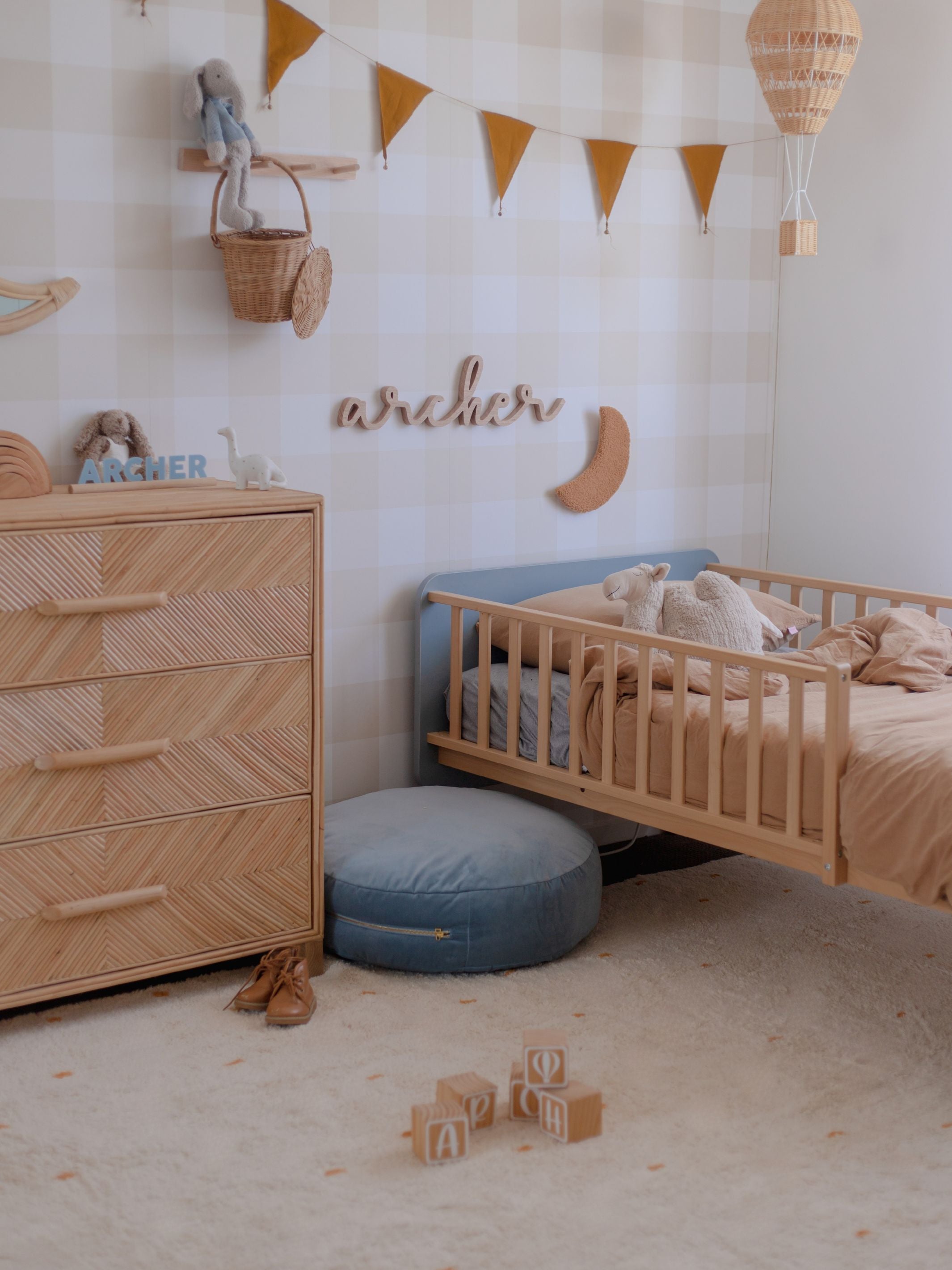 A styled children's bedroom corner featuring a light wood toddler bed with a blue headboard, a matching wooden dresser with woven drawer fronts, a round blue pouf, and warm neutral decor including a hot air balloon wall hanging, bunting, and wooden wall decor.