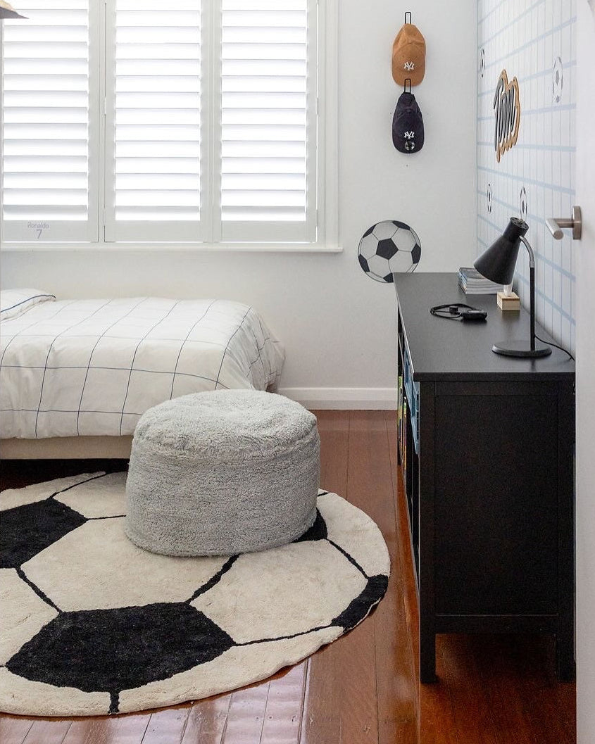 A children's bedroom featuring a soccer-themed decor setup, including a soccer ball patterned rug, a round gray pouf, soccer balls hanging on the wall, and a black dresser against a white paneled wall.